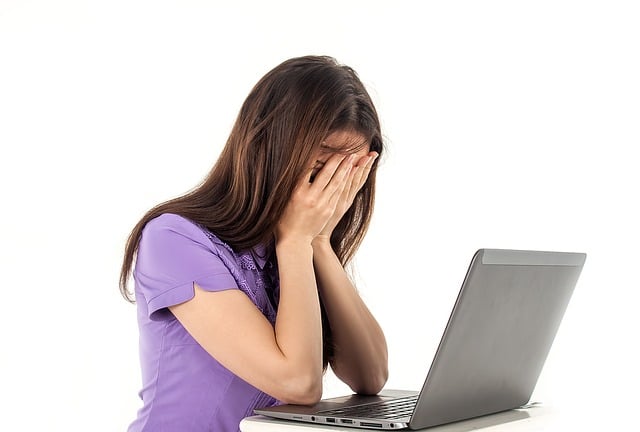 A woman wearing a purple t-shirt sits in front of a laptop computer with her face buried in her hands, looking distraught. She is possibly concerned about the symptoms of Low T in women.