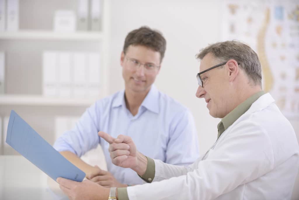 A doctor wearing a white lab coat and glasses discusses with a smiling male patient the results of a low T test at the Prosper clinic.