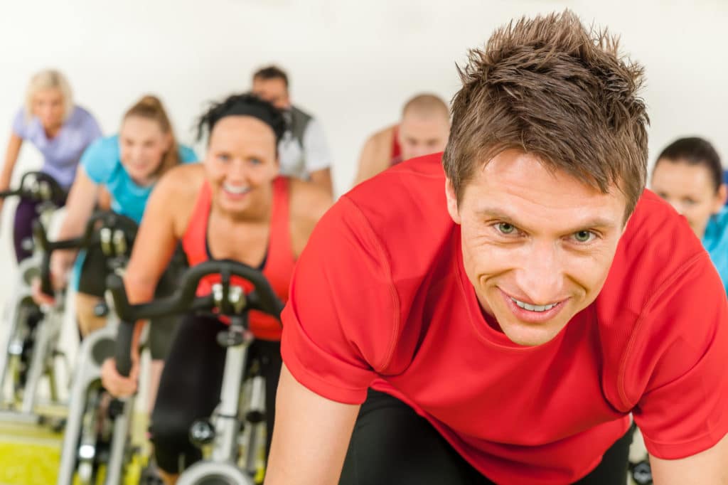 A smiling man in a red t-shirt leads a spin class. Learn about the many exercise trends in Flower Mound.