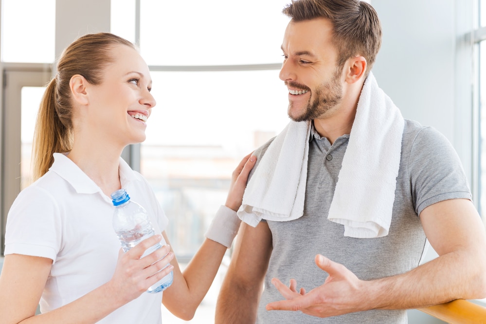 A woman in a white t-shirt talks to a man with short hair, a beard, and a gray t-shirt. The man has a white towel draped around his neck. Both may be members of an employee health initiative in the Dallas-Fort Worth area.