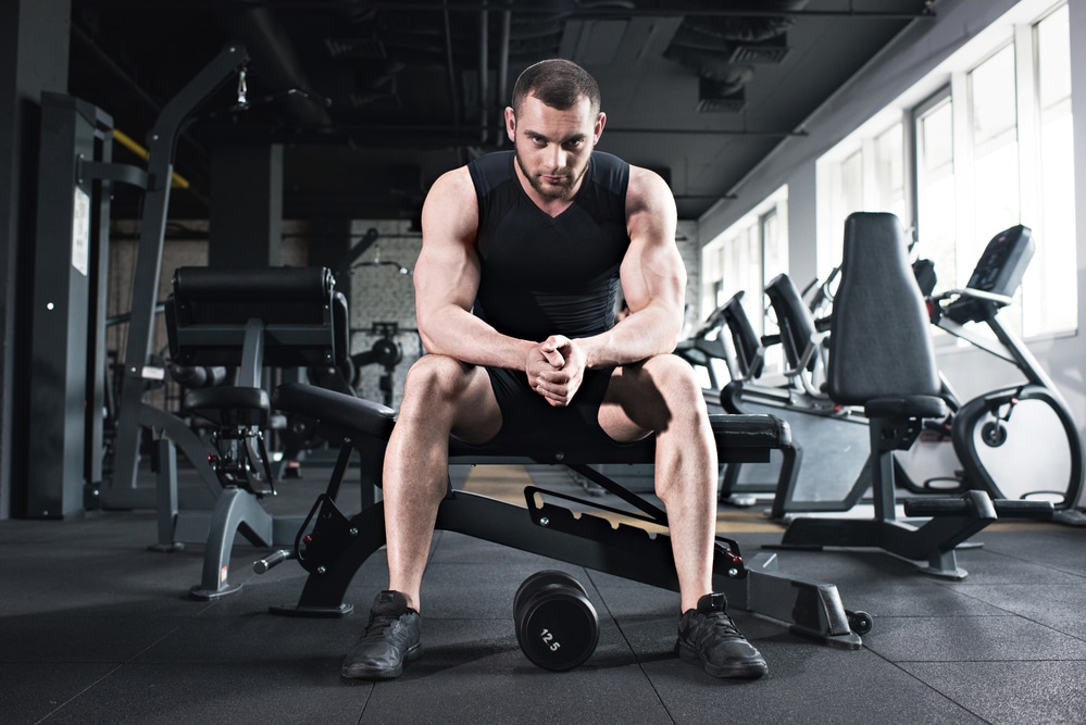A man with short hair wearing a black tank top sits on a weight bench with a dumbbell between his feet. He may be wondering: Is testosterone a steroid?