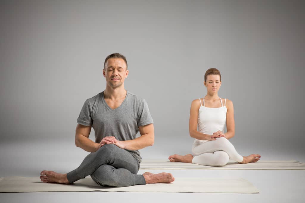A man in gray and a woman in white sit cross-legged as they practice meditation and mindfulness at a DFW studio.