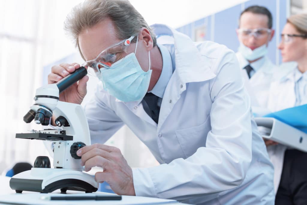 A researcher wearing glasses, mask, and white lab coat looks into a microscope. Scientific research reveals the facts about TRT.