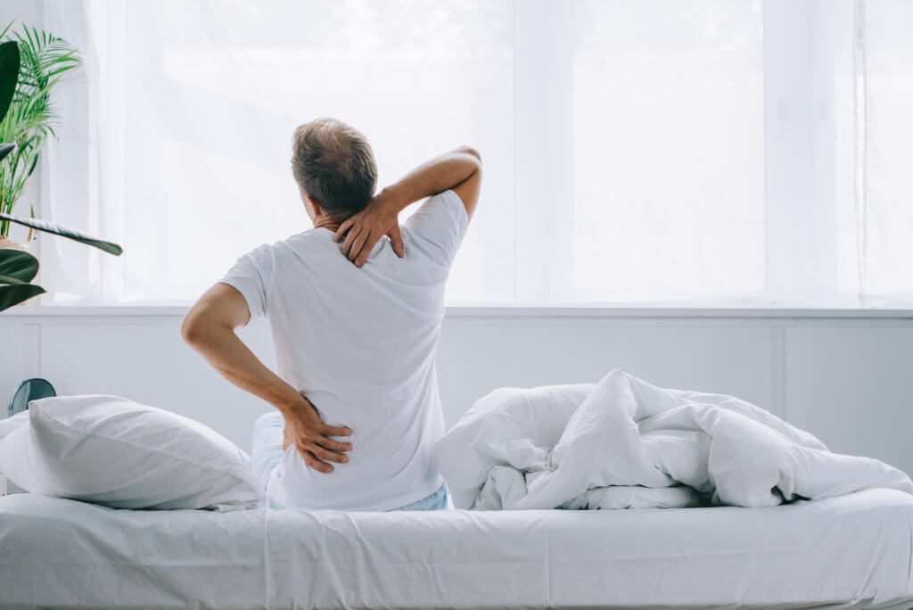 A middle-aged man wearing a white t-shirt sits on a bed with white sheets. He is facing away from the camera, holding his neck and back. Research has found a connection between osteoporosis and low testosterone levels.