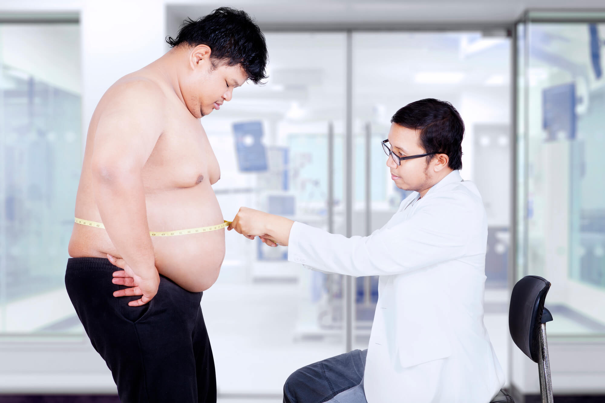 A male doctor uses a measuring tape to measure the stomach of an overweight male patient. The potential role of TRT in the prevention of diabetes may be discussed as part of his treatment plan.