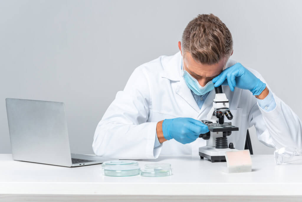 A male scientist in a white lab coat looks down into a microscope while researching the benefits and disadvantages of testosterone pellets.