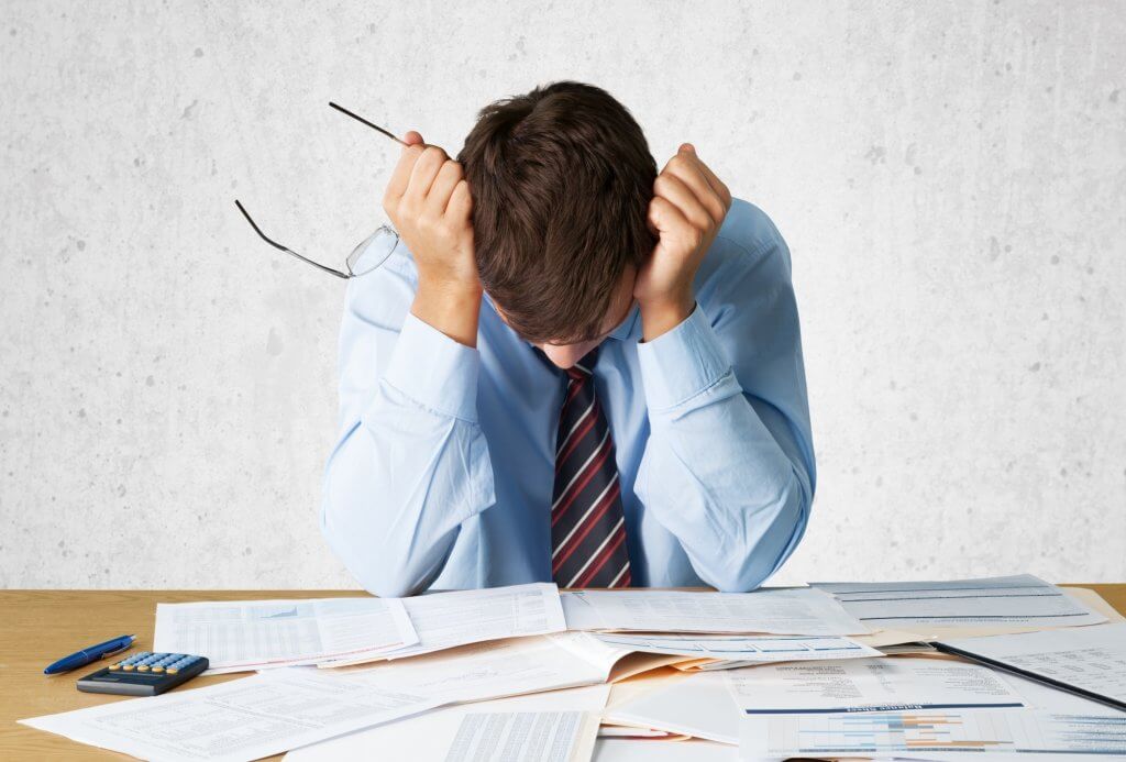 A man sits at a table covered in paper files and charts. He is wearing a blue shirt with a striped tie, and he has his head in his hands. He looks frustrated, possibly due to the connection between hormones and anxiety.