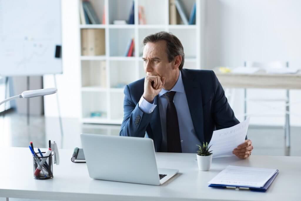 Businessman in a blue suit and tie sits at a white desk and studies reports in front of a white laptop computer. He may be considering the rumors of hGH (human growth hormone) preventing premature aging.