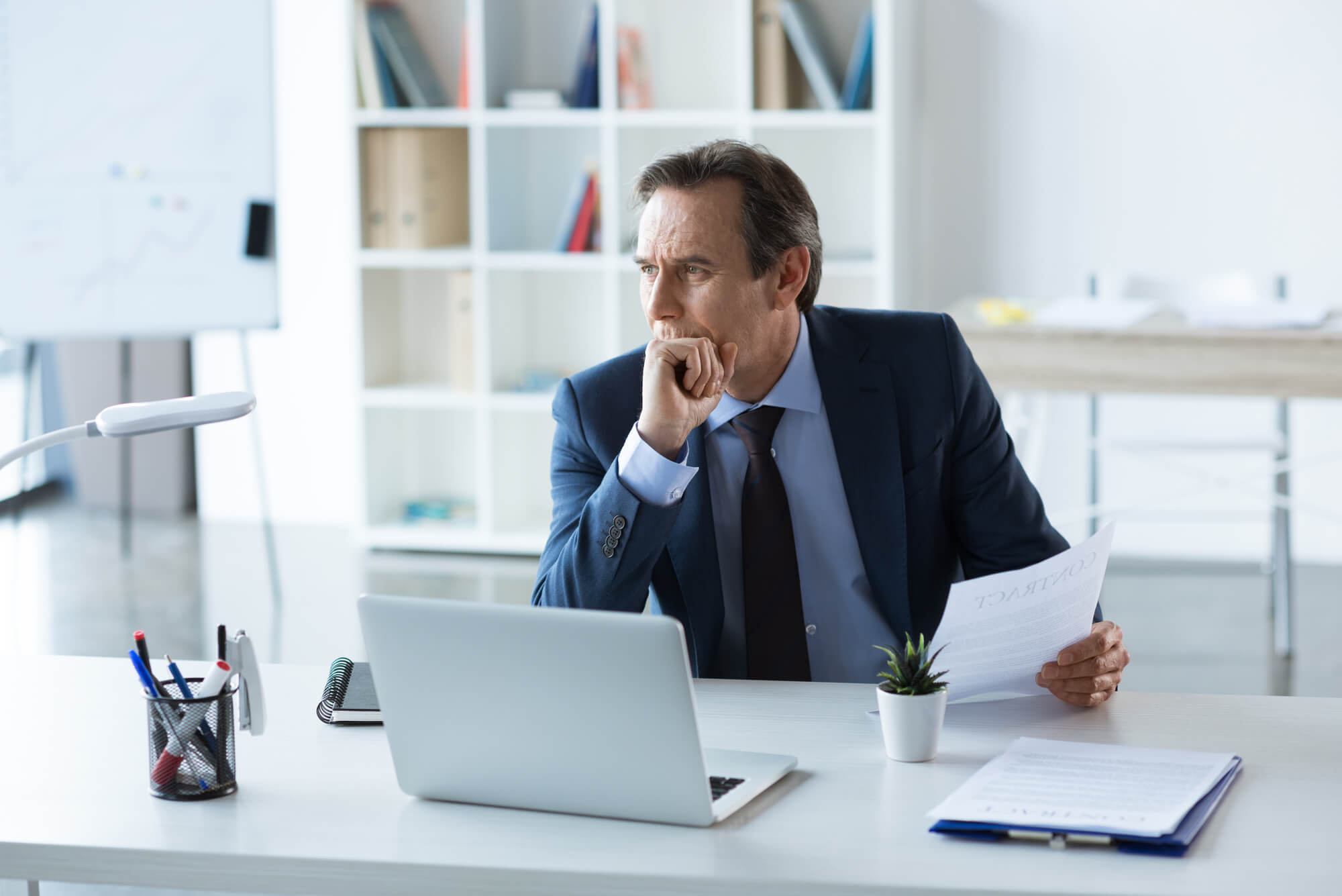 Businessman in a blue suit and tie sits at a white desk and studies reports in front of a white laptop computer. He may be considering the rumors of hGH (human growth hormone) preventing premature aging. 
