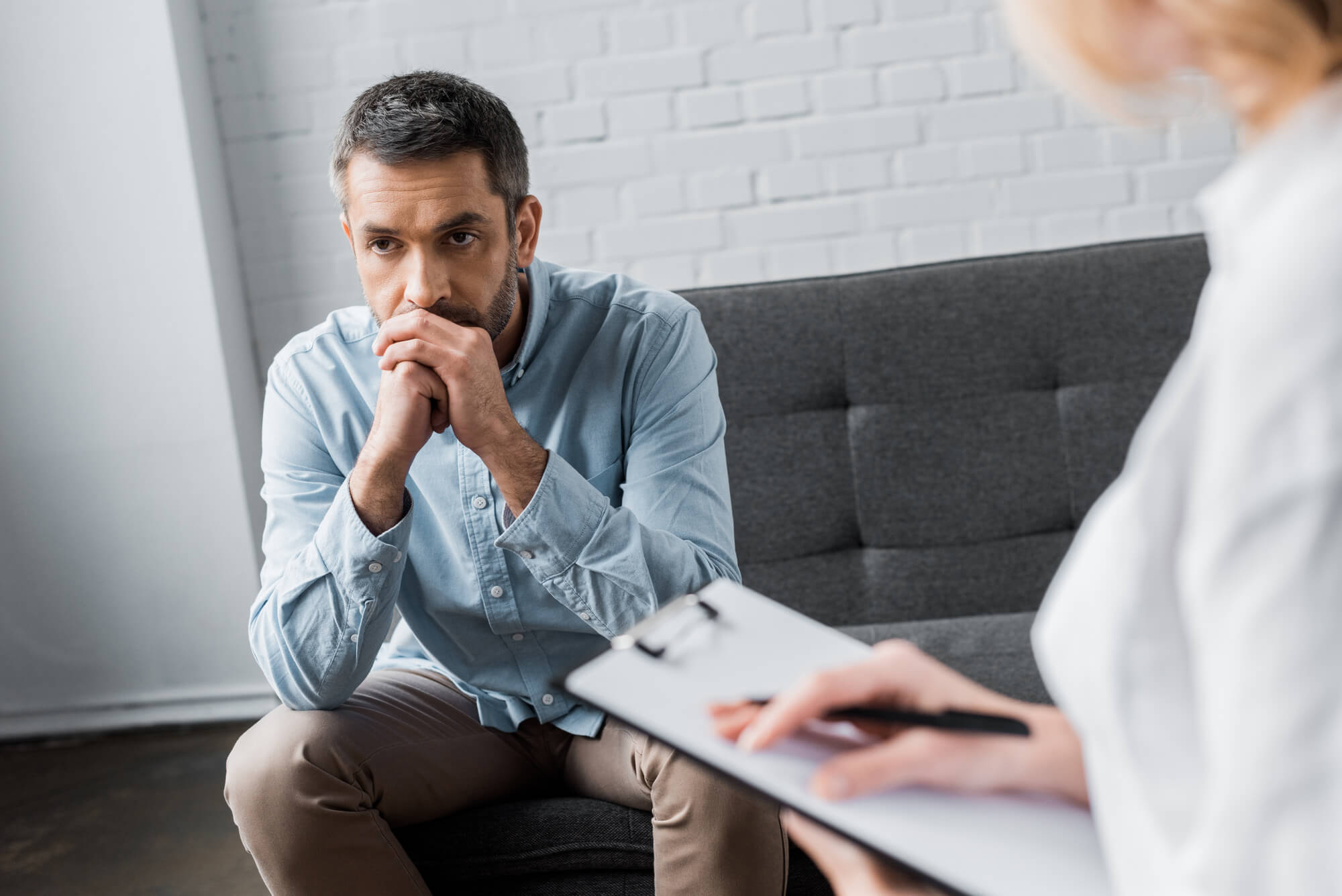 A middle-aged man in a blue shirt sits on a sofa in a doctors office. Doctor is in the foreground, where her hands can be seen holding a clipboard. She may be explaining the options for treating low testosterone with hCG.