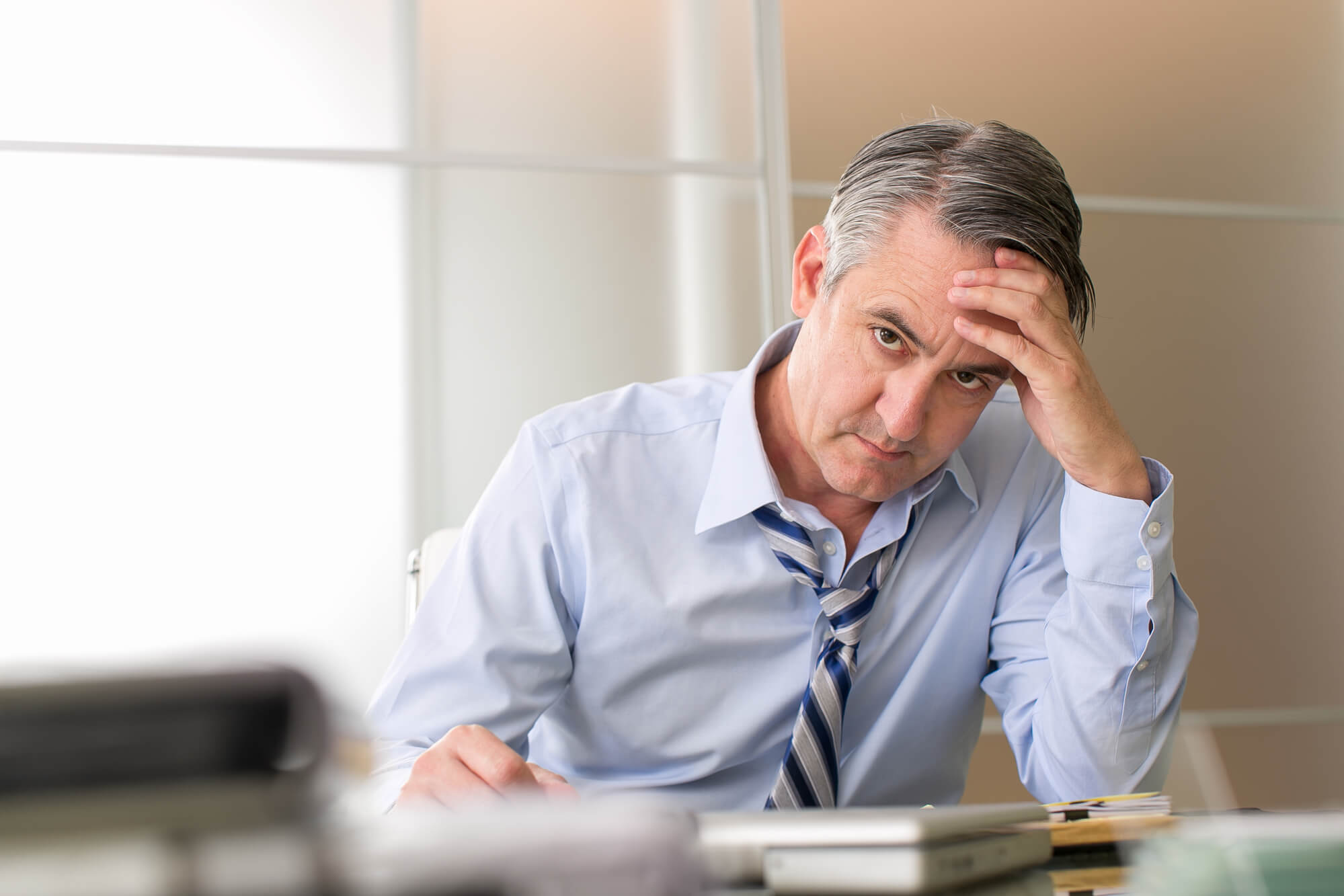 A man in a blue work shirt and striped tie sits at a desk with his head resting on his hand. His high level of stress may be causing low testosterone.
