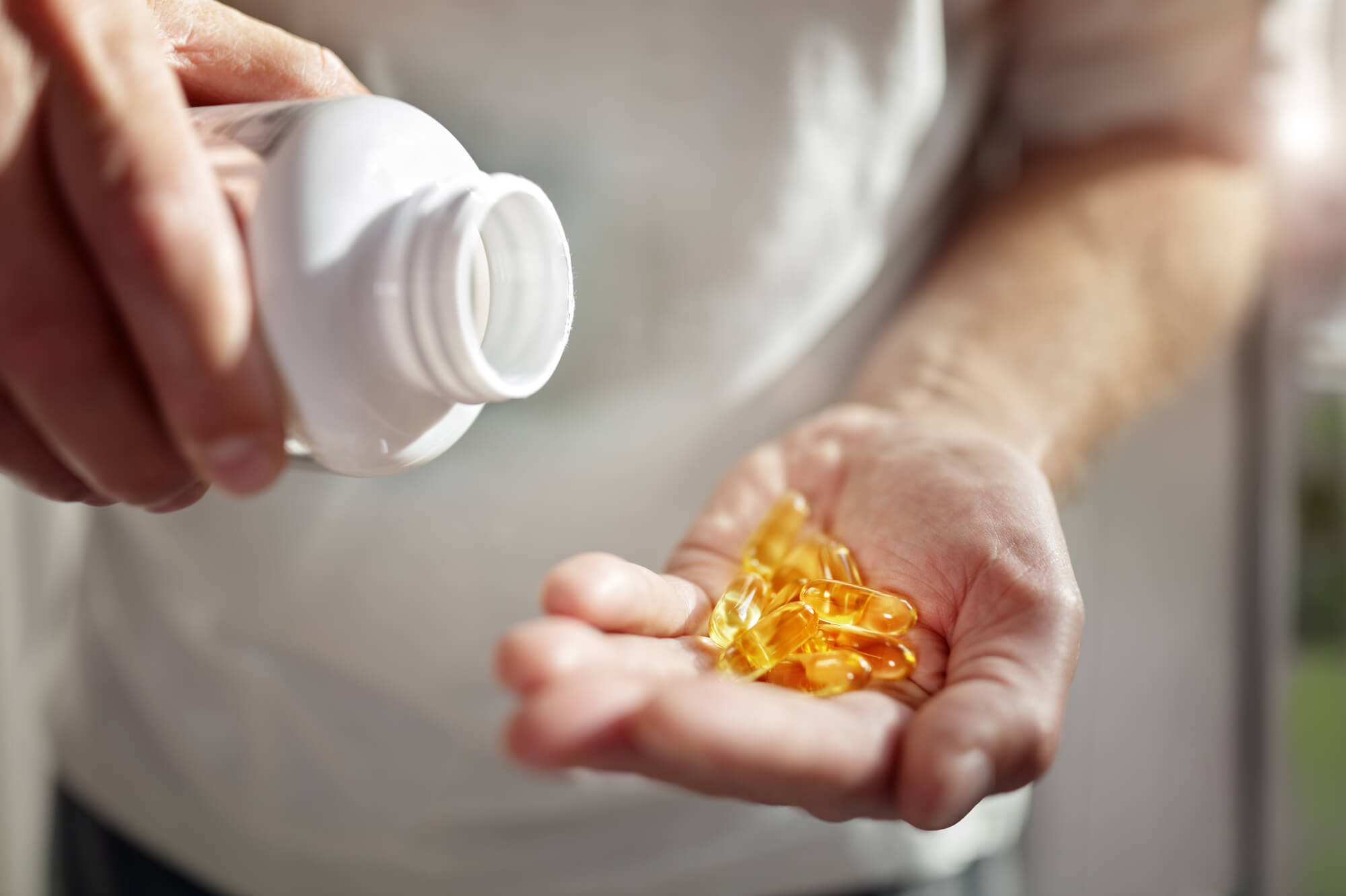 A man in a white t-shirt pours vitamin D gel caps from a white bottle into his cupped hand. Vitamin D may be beneficial to men with low testosterone.