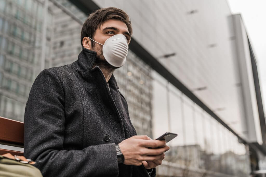 A man in a gray jacket wears a protective face mask as he uses his cellular phone. He may be researching the connection between low testosterone and Covid 19.