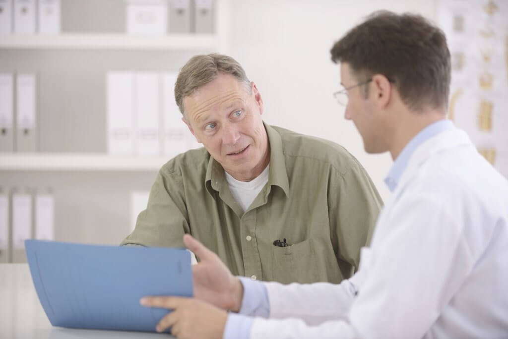 A doctor in a white lab coat talks to a patient wearing a beige button up shirt while pointing to a tablet computer. They may be discussing the possibility of non alcoholic fatty liver disease.
