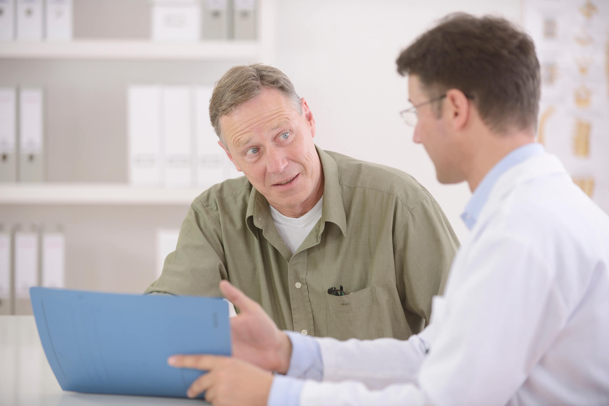A doctor in a white lab coat talks to a patient wearing a beige button up shirt while pointing to a tablet computer. They may be discussing the possibility of non alcoholic fatty liver disease.