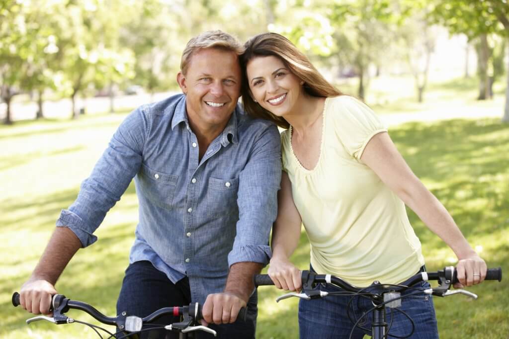 A man in a blue shirt and his female companion ride bikes in the park. Their quality of life has improved since learning the connection between hypothyroidism and low testosterone.