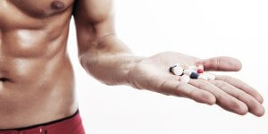 A muscular man's torso with his left arm outstretched can be seen. In his outstretched hand, he is holding a number of pills that could be testosterone boosters.