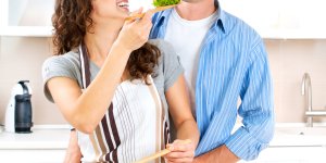 A happy couple are chopping vegetables for a salad. The man stands behind the woman, and she feeds him salad over her left shoulder. He's eating healthy hoping a better diet will help his testosterone.