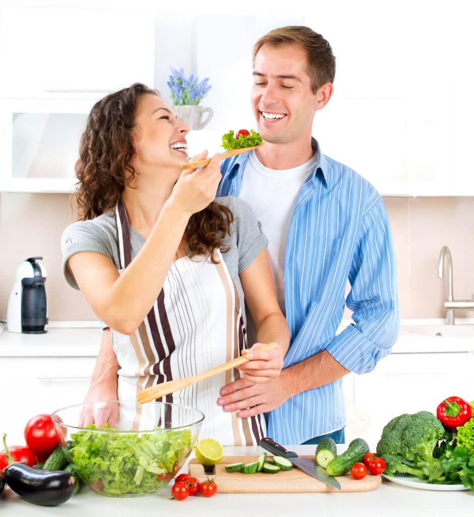 A happy couple are chopping vegetables for a salad. The man stands behind the woman, and she feeds him salad over her left shoulder. He's eating healthy hoping a better diet will help his testosterone.