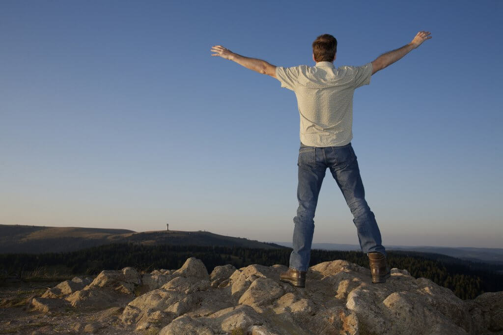 Un hombre se encuentra en la cima de una montaña, mirando una vista del bosque y la puesta de sol en la distancia. Está de espaldas a la cámara y con los brazos abiertos. Como ya no sufre de baja T, sus objetivos de ejercicio eran realistas.