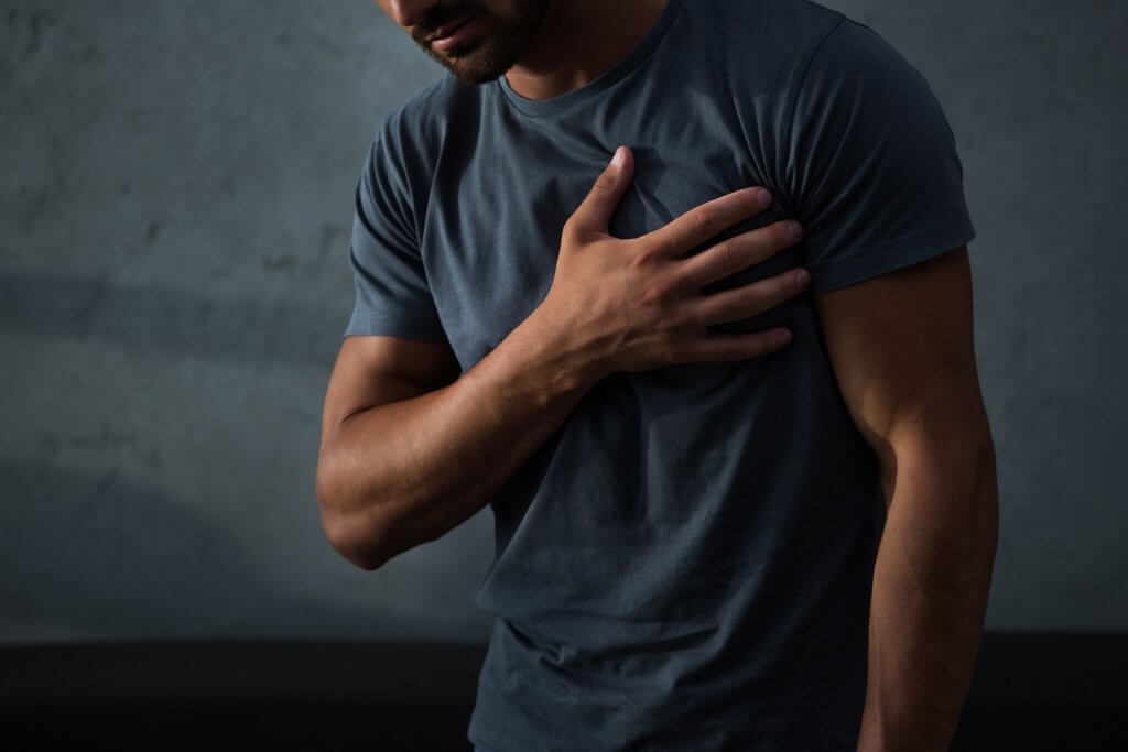 A man in a gray t-shirt clutches his left chest with his right hand. He may be concerned about a heart attack, but research shows this isn't a substantial trt risk.