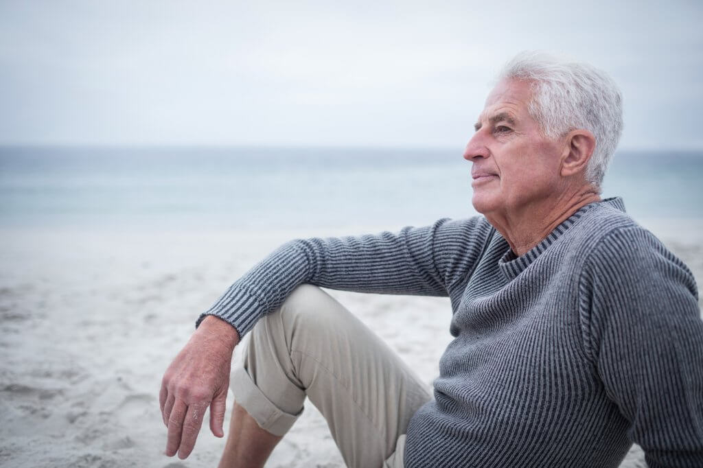 Silver haired man in a gray sweater sits on a beach. He may be suffering the affects of premature aging and low testosterone.