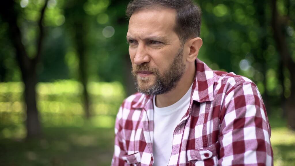 A man with a beard who is wearing a red plaid shirt over a white tee shirt looks concerned, perhaps learning that men need estrogen.