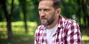A man with a beard who is wearing a red plaid shirt over a white tee shirt looks concerned, perhaps learning that men need estrogen.