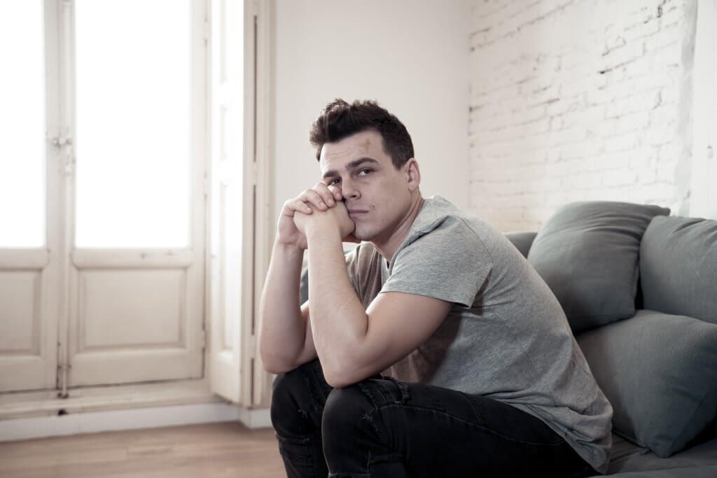 A man in a gray t-shirt sits on the sofa with his head on his hands. He may be feeling the effects of low testosterone and mild depression.