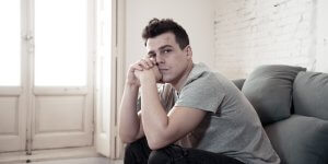 A man in a gray t-shirt sits on the sofa with his head on his hands. He may be feeling the effects of low testosterone and mild depression.