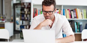 A man wearing a white polo shirt and glasses does research on his laptop, perhaps wondering "What causes low testosterone?"