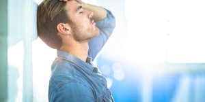 A man in a denim shirt leans back against a shiny wall. He is rubbing his forehead, perhaps worried about chronic Low T Health Risks.