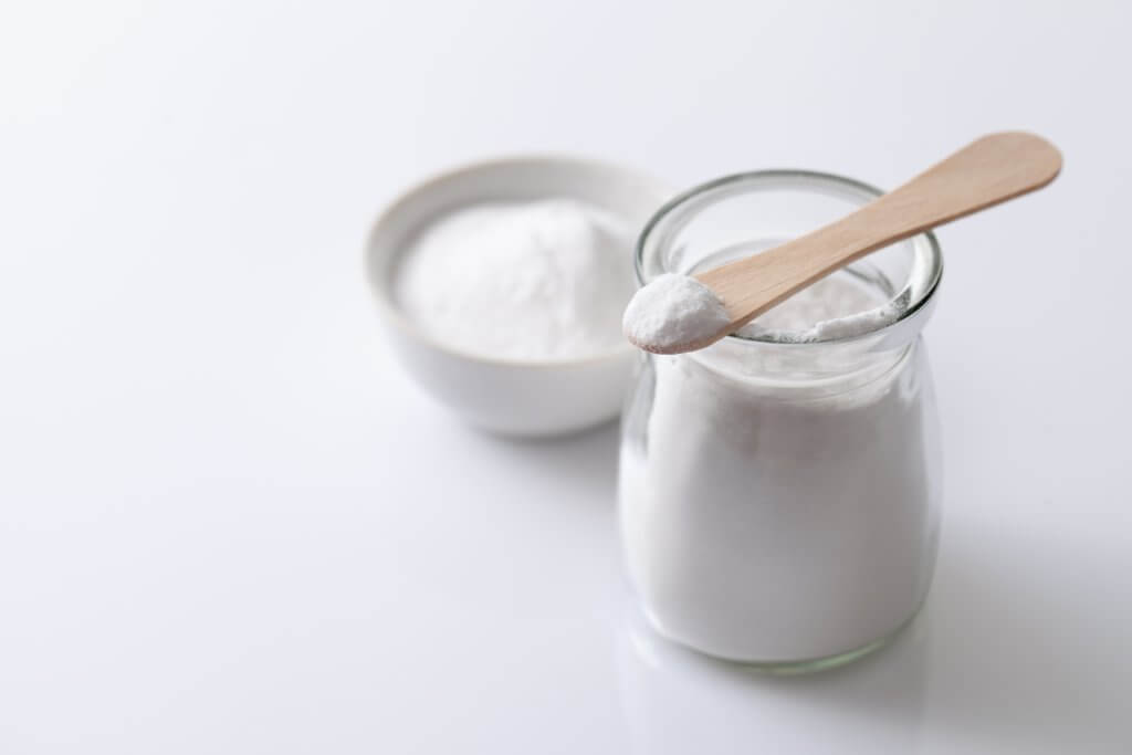 A jar of white powder, possibly sucralose or other artificial sweetener sits on a white surface with a wooden spoon on top.