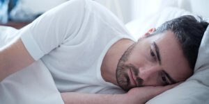 A man in a white t-shirt lies on his side in bed. He may need a sleep apnea mouth guard.
